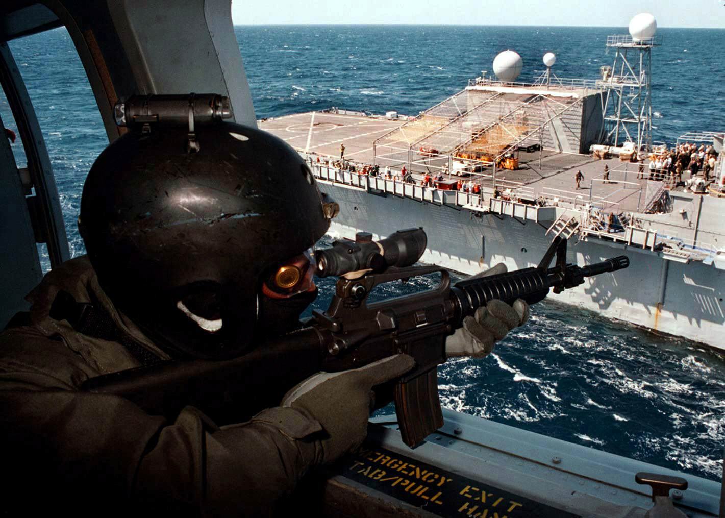 An operator providing overwatch from a helicopter above a naval vessel